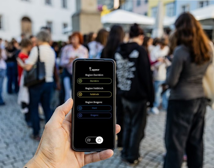 A hand holds a smartphone with the i.appear region selection (Dornbirn, Feldkirch, Hard, Bregenz); in the background, a group of young people on a square in Feldkirch.