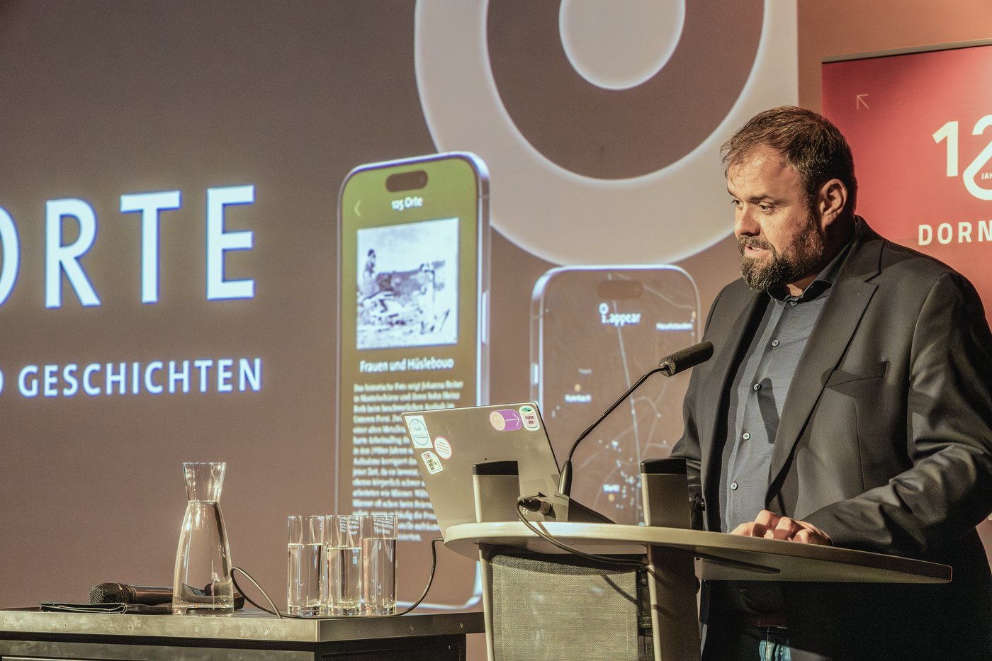 B&uuml;rgermeister Markus F&auml;&szlig;ler h&auml;lt die Er&ouml;ffnungsrede am Rednerpult. Im Hintergrund auf zwei Smartphone-Mockups eine Bildpunkt-Station und die Dornbirn-Karte des Rundgangs.