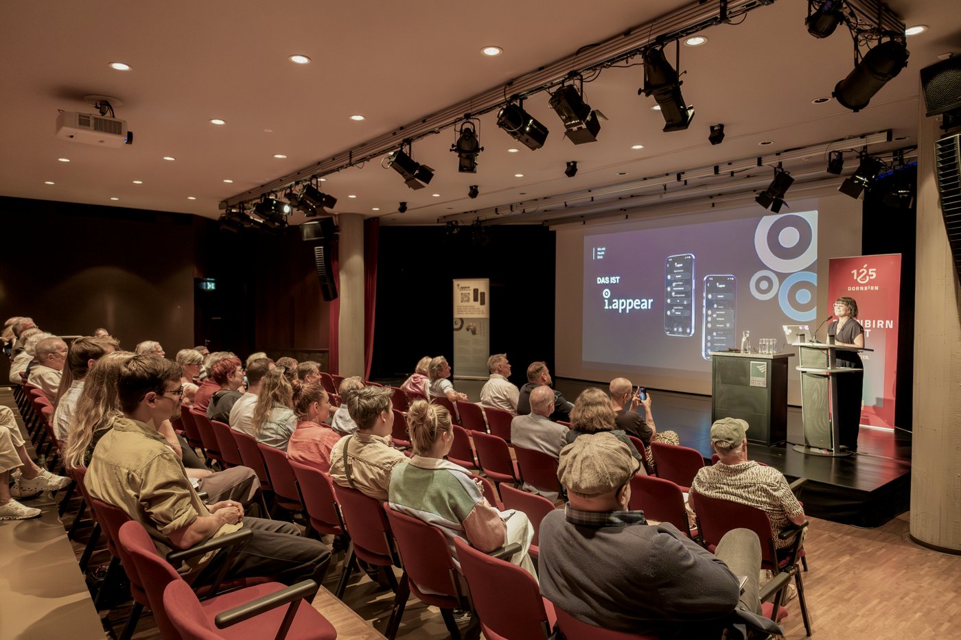 Marilena Tumler pr&auml;sentiert den Rundgang am Rednerpult im Kulturhaus Dornbirn; auf der Leinwand &bdquo;Das ist i.appear&ldquo; mit App-Mockups, davor das Publikum im Saal.